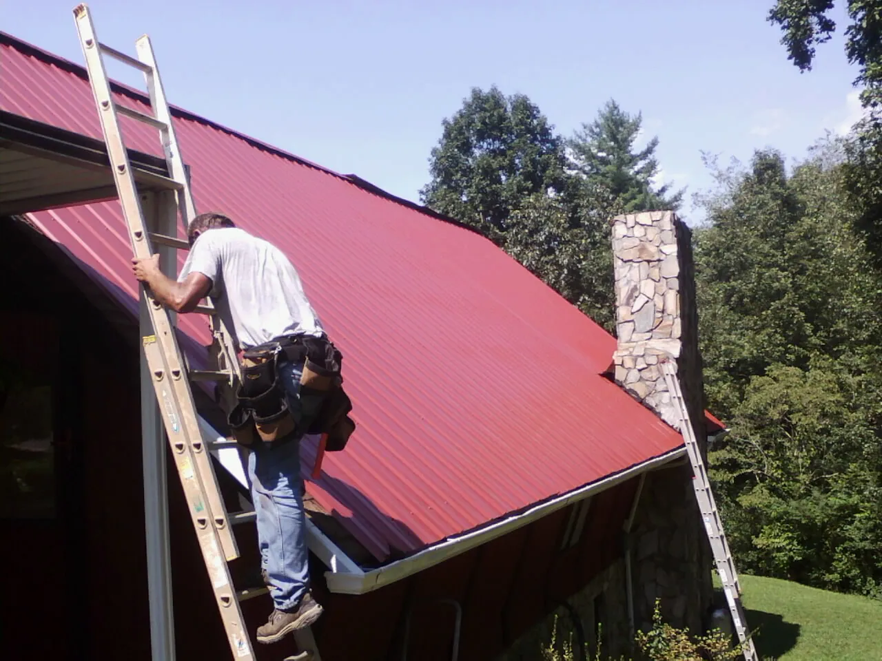 Roofer on a ladder beside a red metal roof and stone chimney — Soffit & Fascia Repair by Roofing Brothers in Hamersville